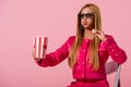 African american woman sitting on chair and eating popcorn isolated on pink, fashion doll concept