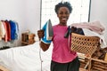 African american woman smiling confident holding basket with clothes and iron machine at bedroom Royalty Free Stock Photo