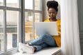 African American woman student sitting on windowsill, working doing remote job on laptop, learning using online course. Self- Royalty Free Stock Photo