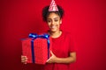 African american woman wearing funny hat holding birthday gift over isolated red background with a happy face standing and smiling Royalty Free Stock Photo