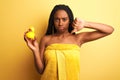 African american woman wearing shower towel holding toy duck over isolated yellow background with angry face, negative sign