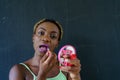 African American woman with yellow braids holding small mirror and wearing purple lipstick while looking at the camera Royalty Free Stock Photo