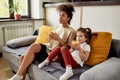 Enjoy the day. African american woman baby sitter entertaining caucasian cute little girl. They are sitting on the couch Royalty Free Stock Photo