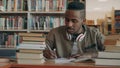 Positive african american young handsome guy with big headphones is sitting at table with books and writing in his Royalty Free Stock Photo