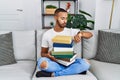 African american young man holding a pile of books sitting on the sofa checking the time on wrist watch, relaxed and confident