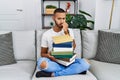 African american young man holding a pile of books sitting on the sofa looking stressed and nervous with hands on mouth biting Royalty Free Stock Photo