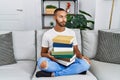 African american young man holding a pile of books sitting on the sofa smiling looking to the side and staring away thinking Royalty Free Stock Photo