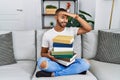 African american young man holding a pile of books sitting on the sofa very happy and smiling looking far away with hand over head Royalty Free Stock Photo