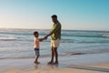 African american young man holding son's hands while standing at beach against clear sky at sunset Royalty Free Stock Photo