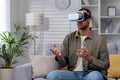 African american young man sitting on sofa at home wearing 3d glasses, wearing virtual reality glasses, playing online Royalty Free Stock Photo