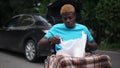 African American young man in wheelchair with groceries outdoors with blurred black car at background. Portrait of sad