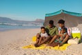 African american young parents with children sitting under umbrella on blanket at beach against sky Royalty Free Stock Photo