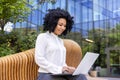 African American young smiling businesswoman sitting on a bench outside an office center and working remotely on a Royalty Free Stock Photo