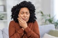 African american young woman sitting on sofa at home and holding hands to cheek, eyes closed, suffering from toothache Royalty Free Stock Photo