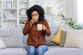 African american young woman wiping nose with tissue from cold, sitting on sofa at home sick and holding cup in hands Royalty Free Stock Photo