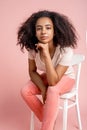 Freestyle. African girl sitting on chair isolated on pink leaning on hand smiling pensive close-up Royalty Free Stock Photo