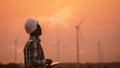 African engineer windmills wearing white hard hat and using tablet with wind turbine on silhouette sunset