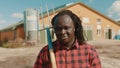 African farmer holding fork over the soulder and nodding his head in front of the silo system