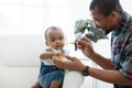 African father feeding adorable toddler baby girl with spoon while sitting on sofa at home and kid`s face mess up with food.