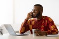 African Freelancer Guy Talking On Cellphone And Enjoying Coffee At Home Office Royalty Free Stock Photo