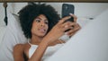 African girl relaxing cellphone smiling bedroom. Curly lady laying soft pillow