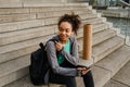 African girl using mobile phone while sitting with yoga mat on stairs Royalty Free Stock Photo