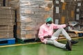 African male inventory control manager in sterile uniform sitting and checking order of cardboard box package stacked in warehouse