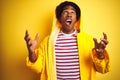 African man with afro hair wearing rain coat with hood standing over isolated yellow background crazy and mad shouting and yelling