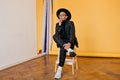 African man in white sneakers sitting on chair in studio. Indoor photo of pensive mulatto guy wears trendy dark-gray