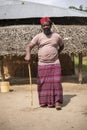An African Older Man in Red Muslim Taqiyyah Fez Hat posing with a stick for lame people on Yard Near the Basic Hut with