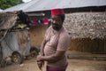 An African Older Man in Red Muslim Taqiyyah Fez Hat posing with a stick for lame people on Yard Near the Basic Hut with