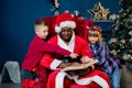 African Santa Claus is sitting on a chair and reading a book with Christmas stories for two young children