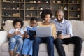 African couple with kids sit on sofa with diverse gadgets