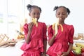 African twin girl sister with curly hair braid African hairstyle eating banana in kitchen. Happy smiling kid sibling eating fruit Royalty Free Stock Photo