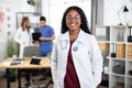 African woman doctor holding hands in pockets of scrubs and smiling, standing in modern clinic room Royalty Free Stock Photo