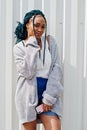 African woman with dreads hair, in jeans shorts  posed against white steel wall with mobile phone in hand Royalty Free Stock Photo