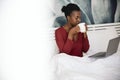 African woman sitting in bed sipping on coffee looking at laptop Royalty Free Stock Photo