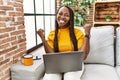 African woman sitting on the sofa using laptop at home very happy and excited doing winner gesture with arms raised, smiling and Royalty Free Stock Photo