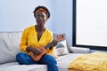 African young woman playing ukulele at home afraid and shocked with surprise and amazed expression, fear and excited face