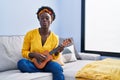 African young woman playing ukulele at home making fish face with mouth and squinting eyes, crazy and comical