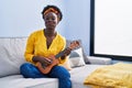 African young woman playing ukulele at home puffing cheeks with funny face