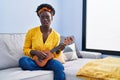 African young woman playing ukulele at home in shock face, looking skeptical and sarcastic, surprised with open mouth