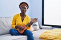 African young woman playing ukulele at home winking looking at the camera with sexy expression, cheerful and happy face
