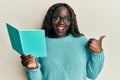 African young woman reading a book wearing glasses pointing thumb up to the side smiling happy with open mouth Royalty Free Stock Photo