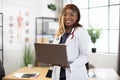 Afro-American doctor wearing white coat with stethoscope, working on laptop pc in hospital office Royalty Free Stock Photo