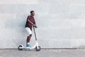 Afro american guy rides an electric scooter against the background of a wall, student uses eco transport