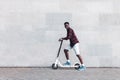 Afro american guy rides an electric scooter against the background of a wall, student uses eco transport