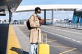 Afro-American millennial traveler man with suitcase stands in airport terminal, holding mobile phone, calling for a taxi or car Royalty Free Stock Photo