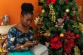 Afro-descendant Colombian woman sitting on the sofa next to the Christmas tree with her cell phone