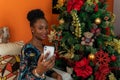 Afro-descendant Colombian woman sitting on the sofa next to the Christmas tree with her cell phone